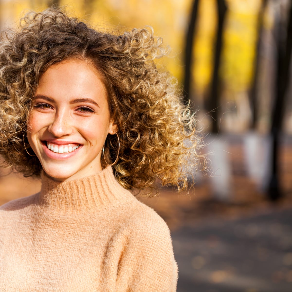 A smiling woman with curly blonde hair outdoors in autumn light, beside the review she left for the practice