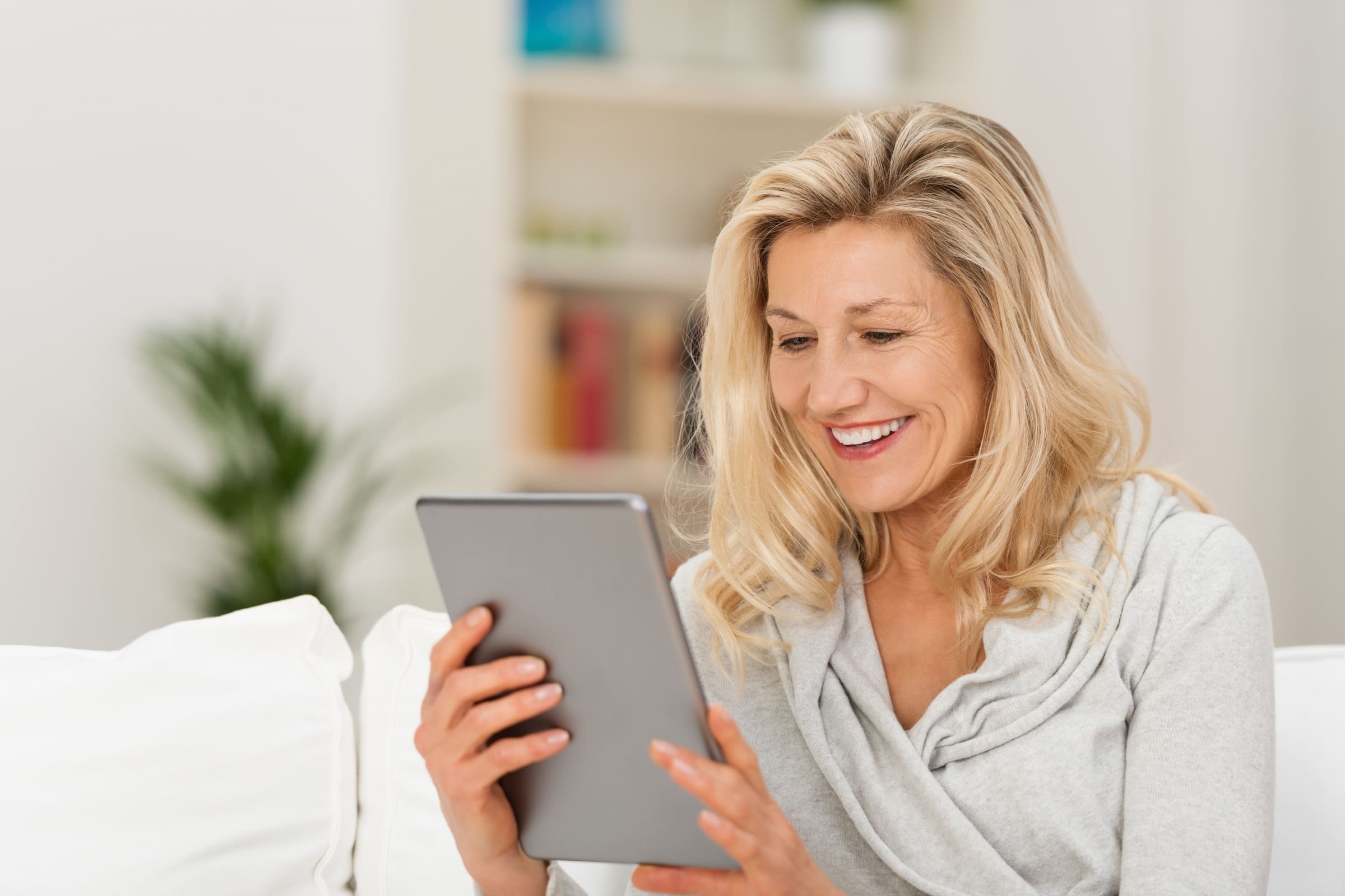 A smiling woman with blonde hair reading a tablet on a sofa at home
