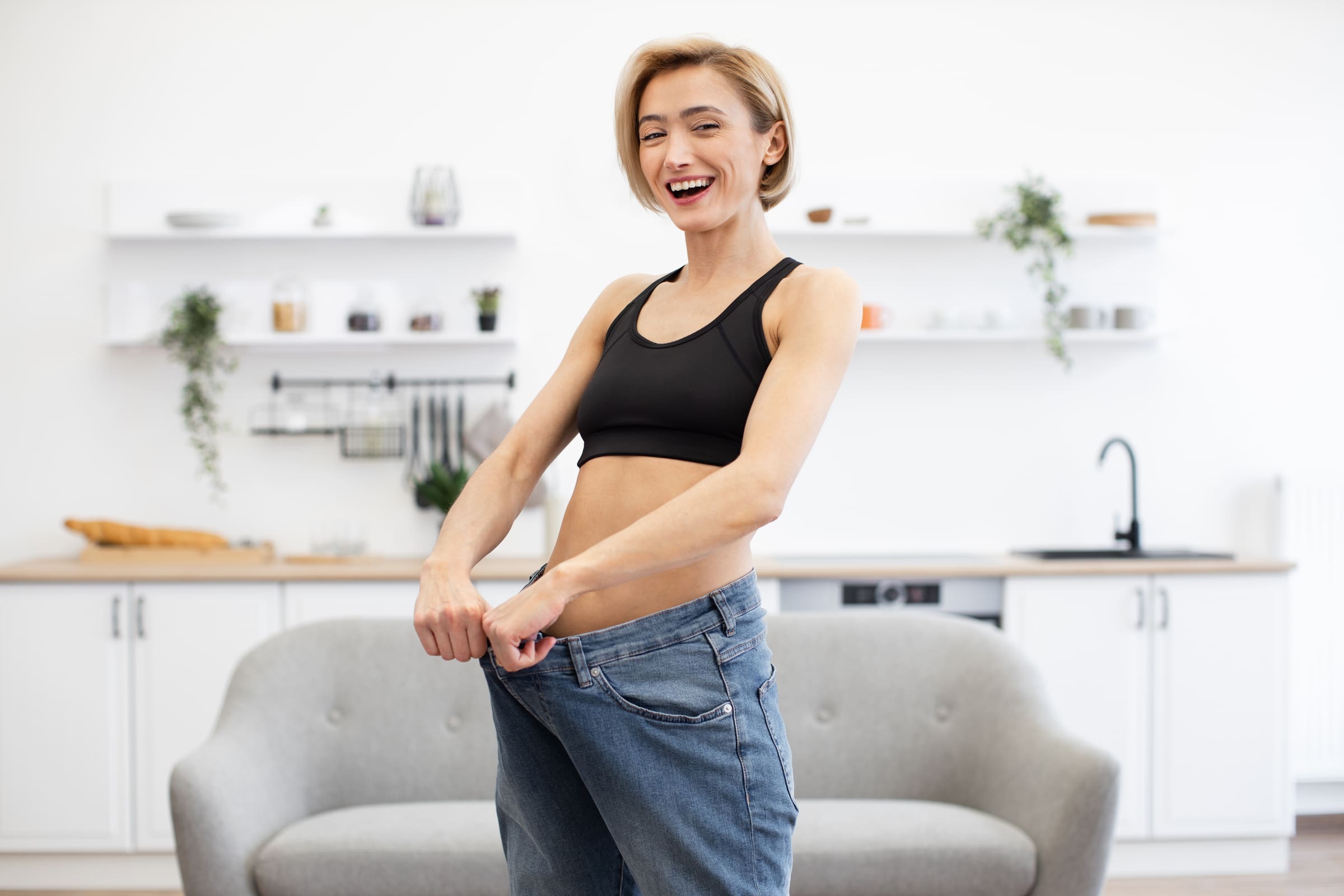 A smiling woman in a black sports top holding out the waistband of jeans now far too big for her, in a bright kitchen