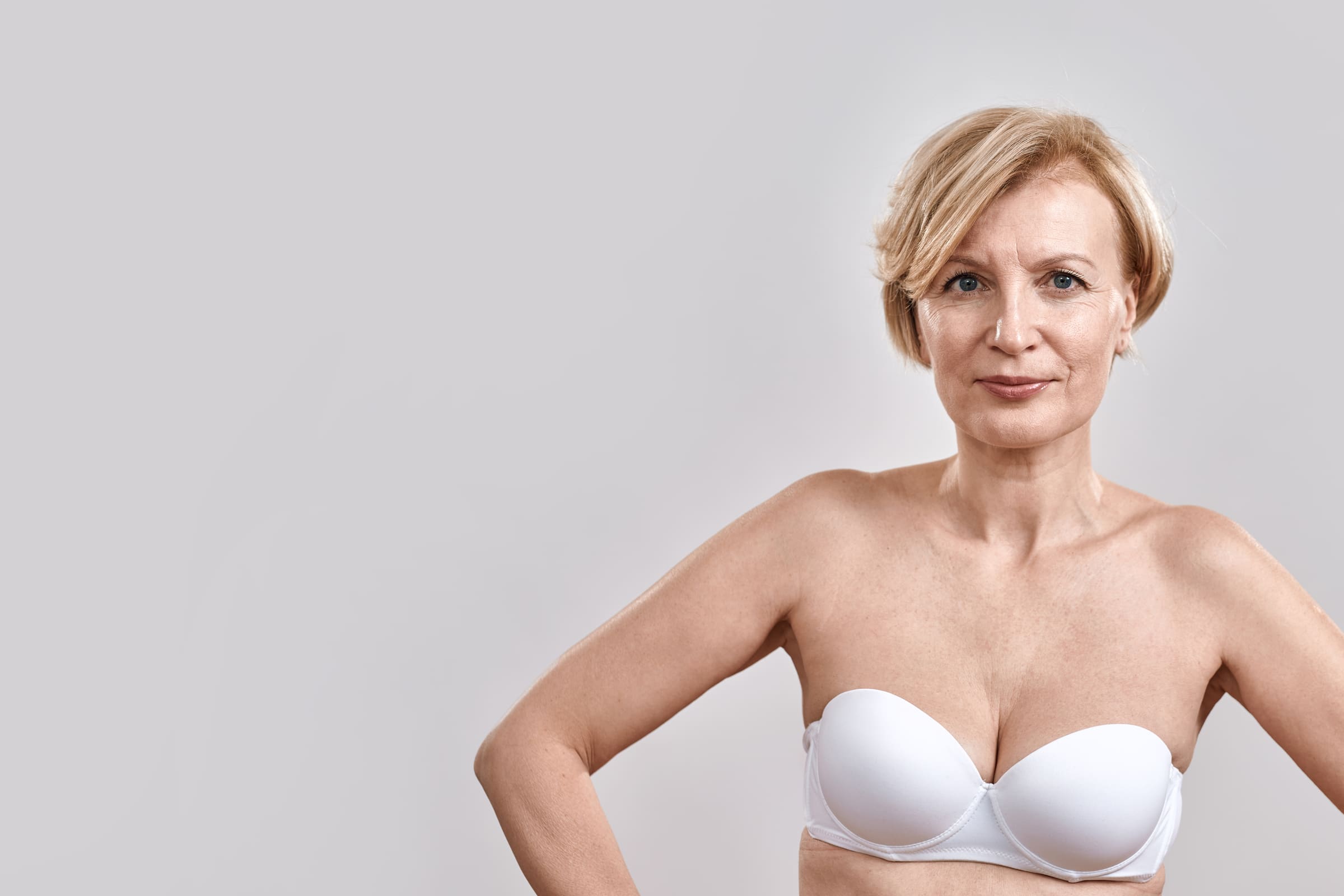 A confident middle-aged woman with short blonde hair in a white bra, hands on her hips, against a grey background