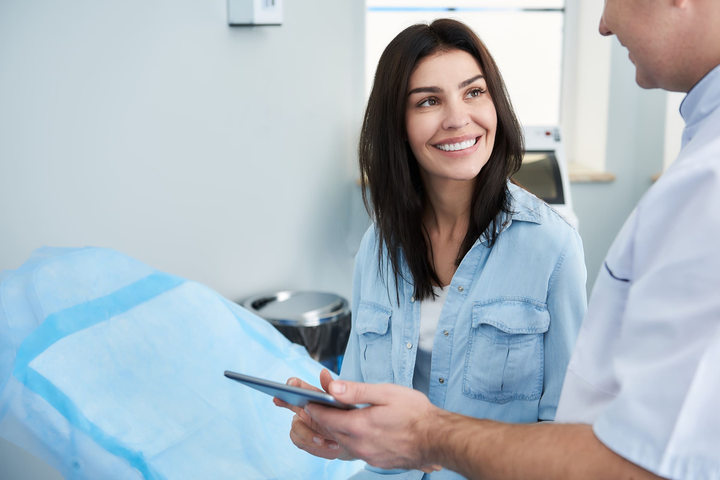 A smiling woman holding a tablet while a plastic surgeon in a white coat talks her through it in a bright clinic