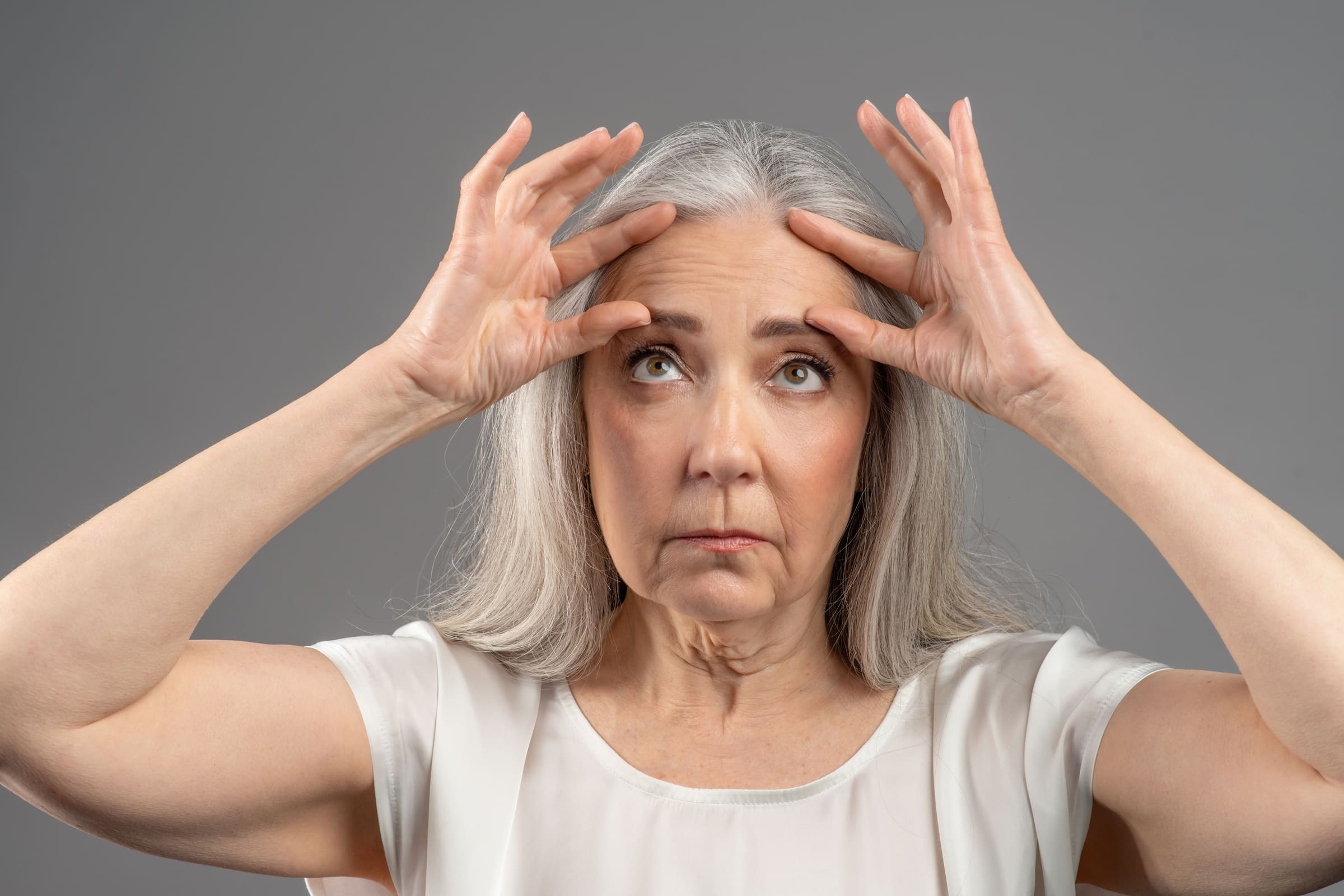 A mature woman with long grey hair gently lifting the skin of her forehead with her fingertips