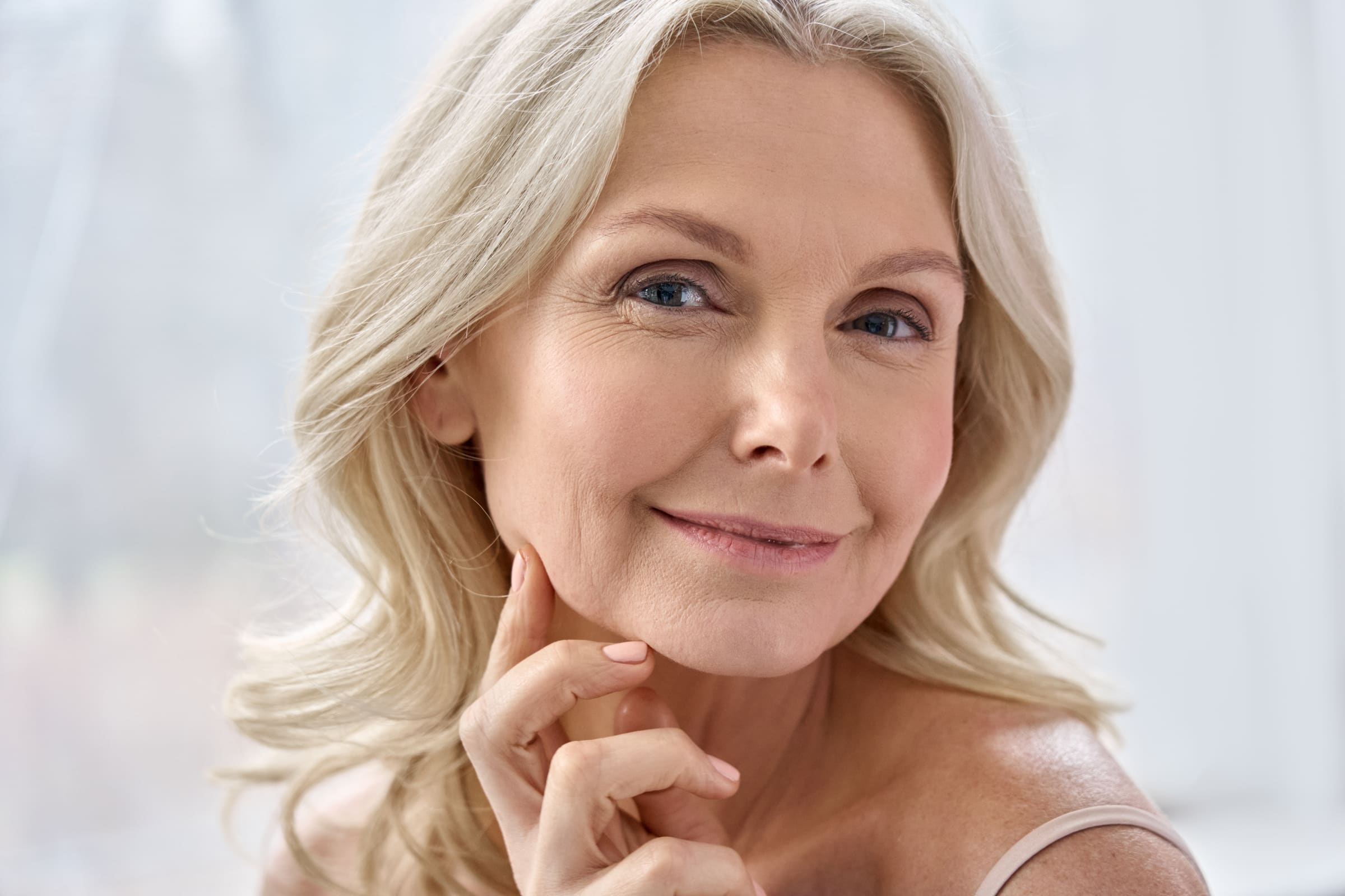 A smiling mature woman with blonde hair resting her fingers on her chin in soft daylight