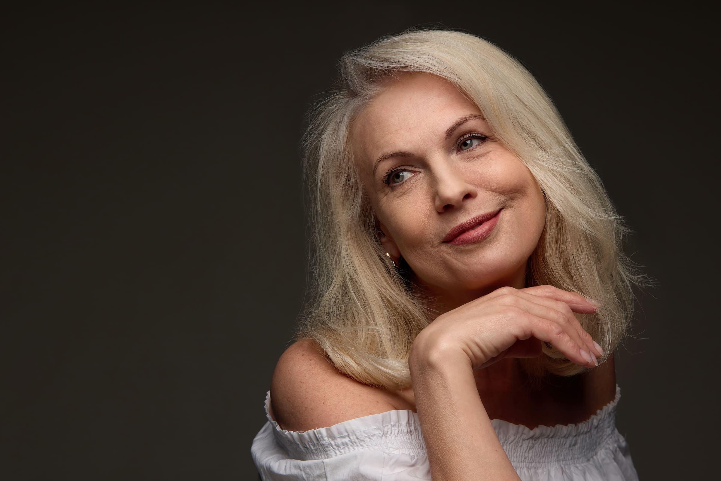 A smiling mature woman with blonde hair resting her chin on her hand against a dark background