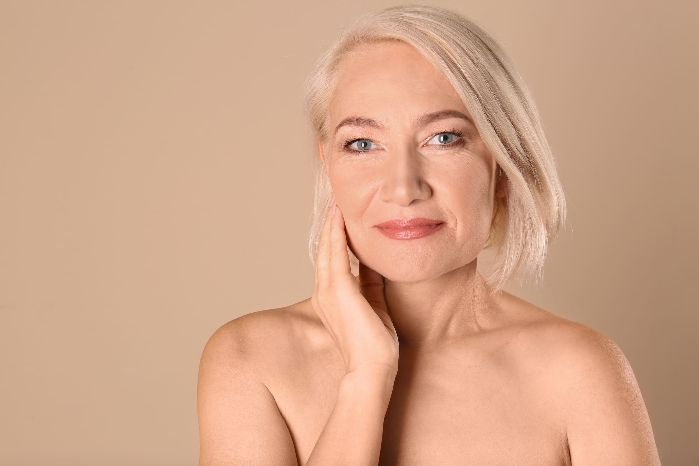 A mature woman with short blonde hair touching her jawline against a warm beige background