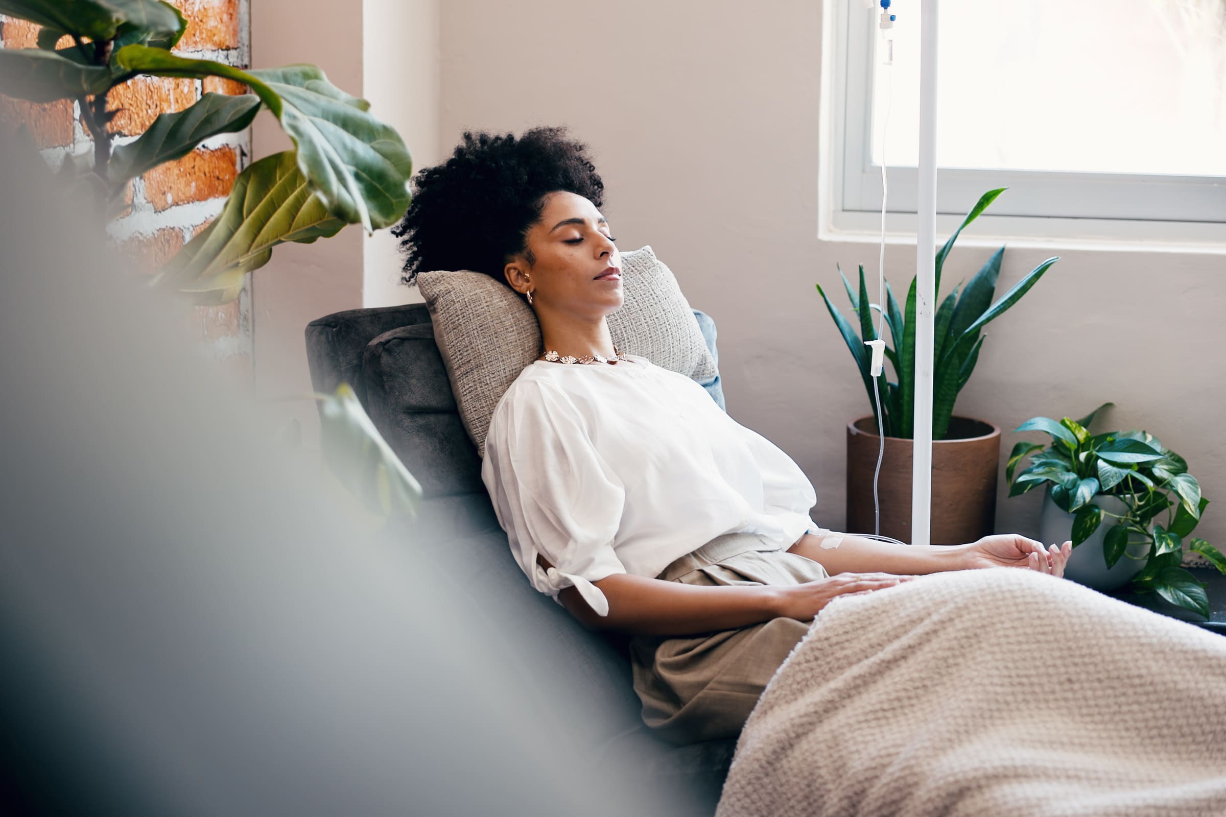 A woman relaxing in a recliner with her eyes closed while receiving an IV drip in a bright, plant-filled lounge