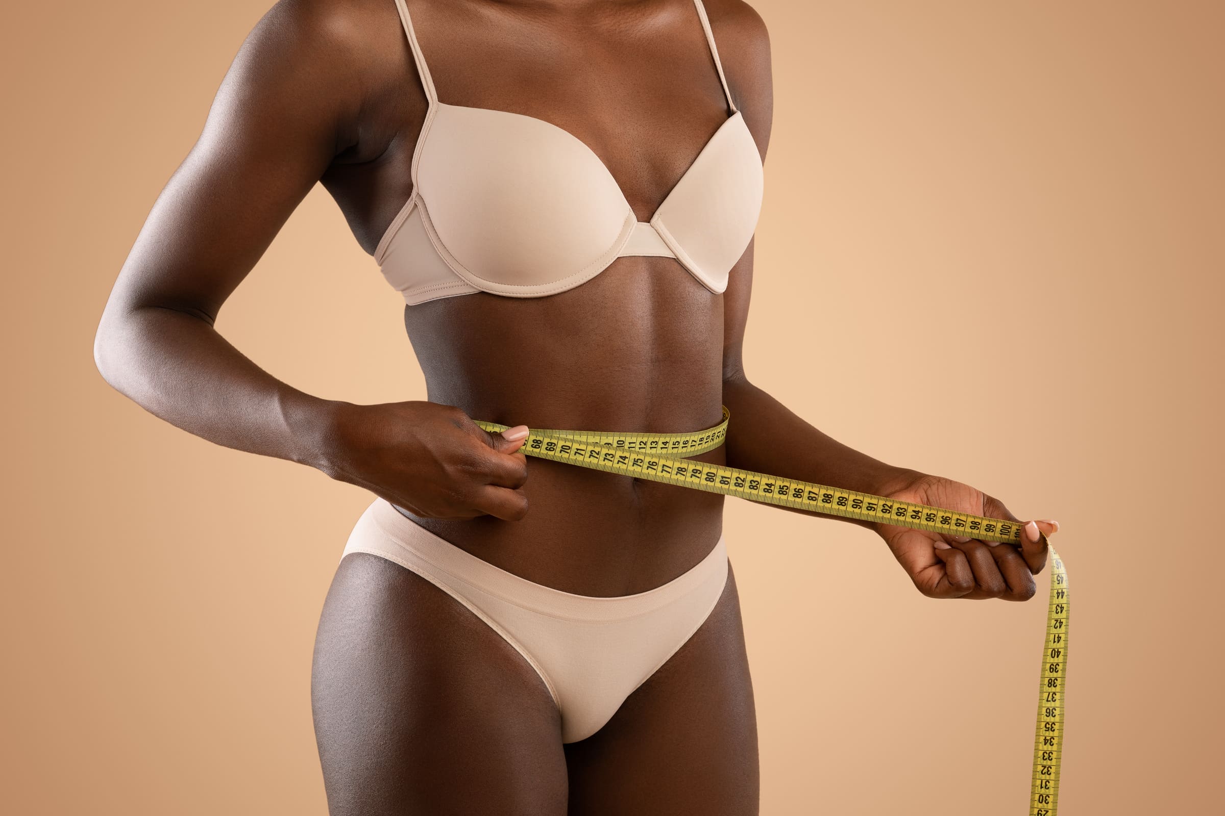 A woman in beige lingerie measuring her waist with a yellow tape measure against a warm beige background