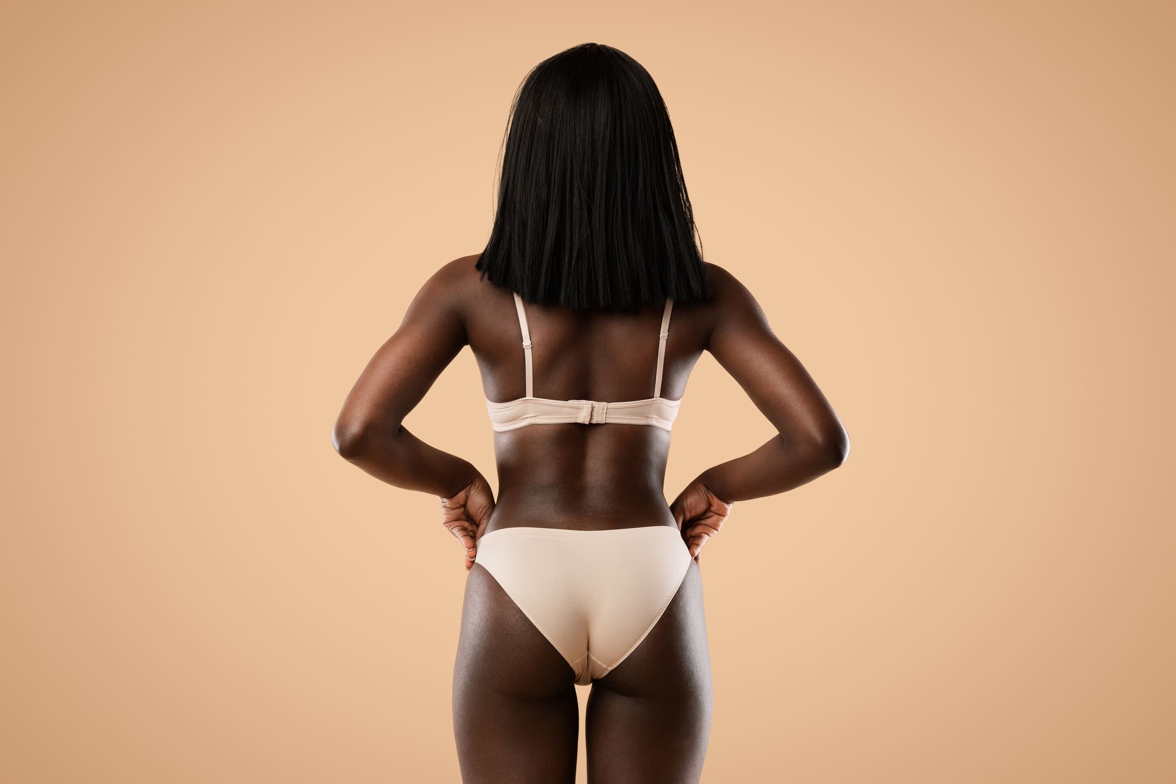 A woman in beige underwear seen from behind, hands on her hips, against a warm beige background