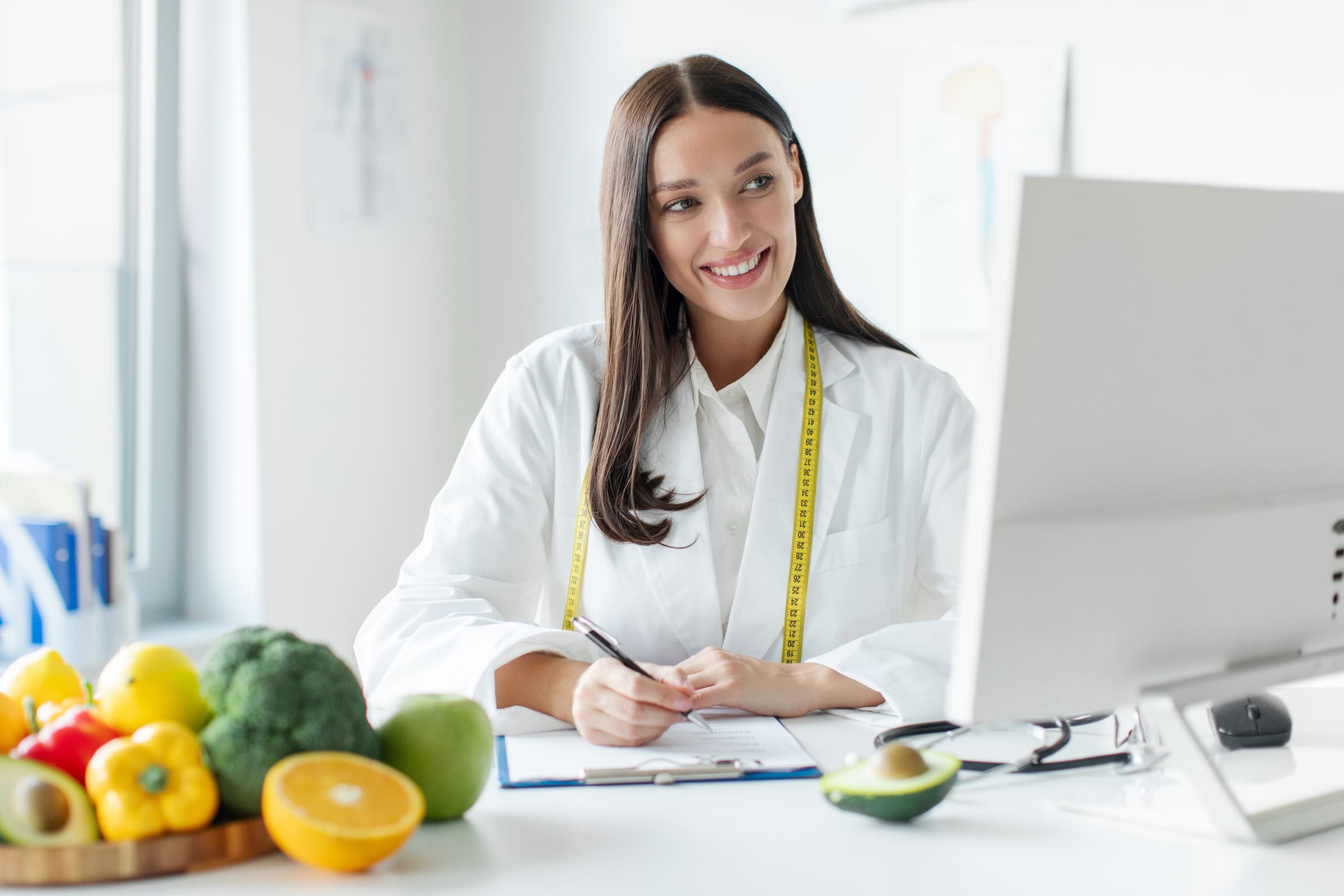 A smiling doctor with a tape measure around her neck writing at a desk beside a bowl of fresh fruit and vegetables