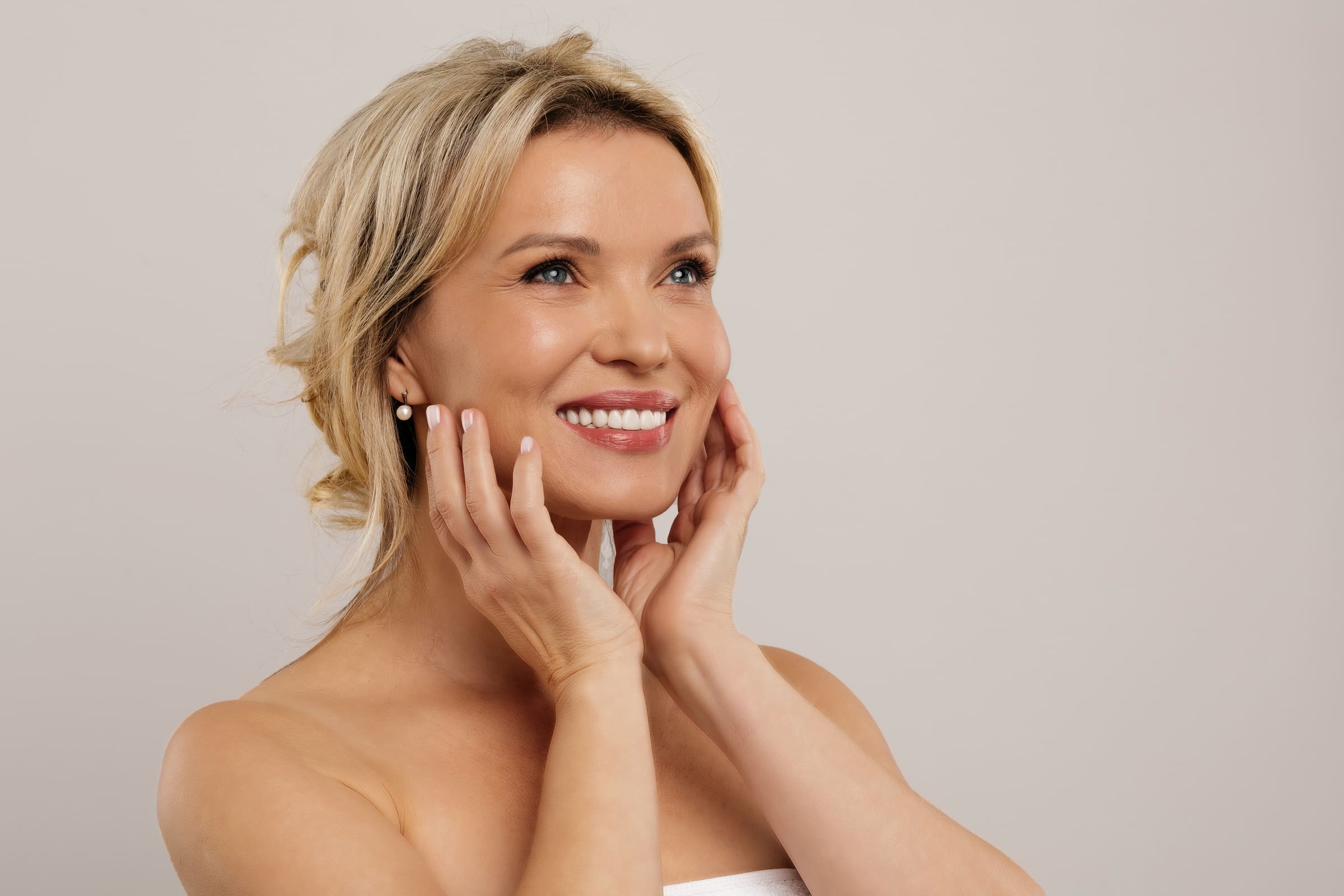 A smiling woman with short blonde hair cradling her face in her hands against a neutral background