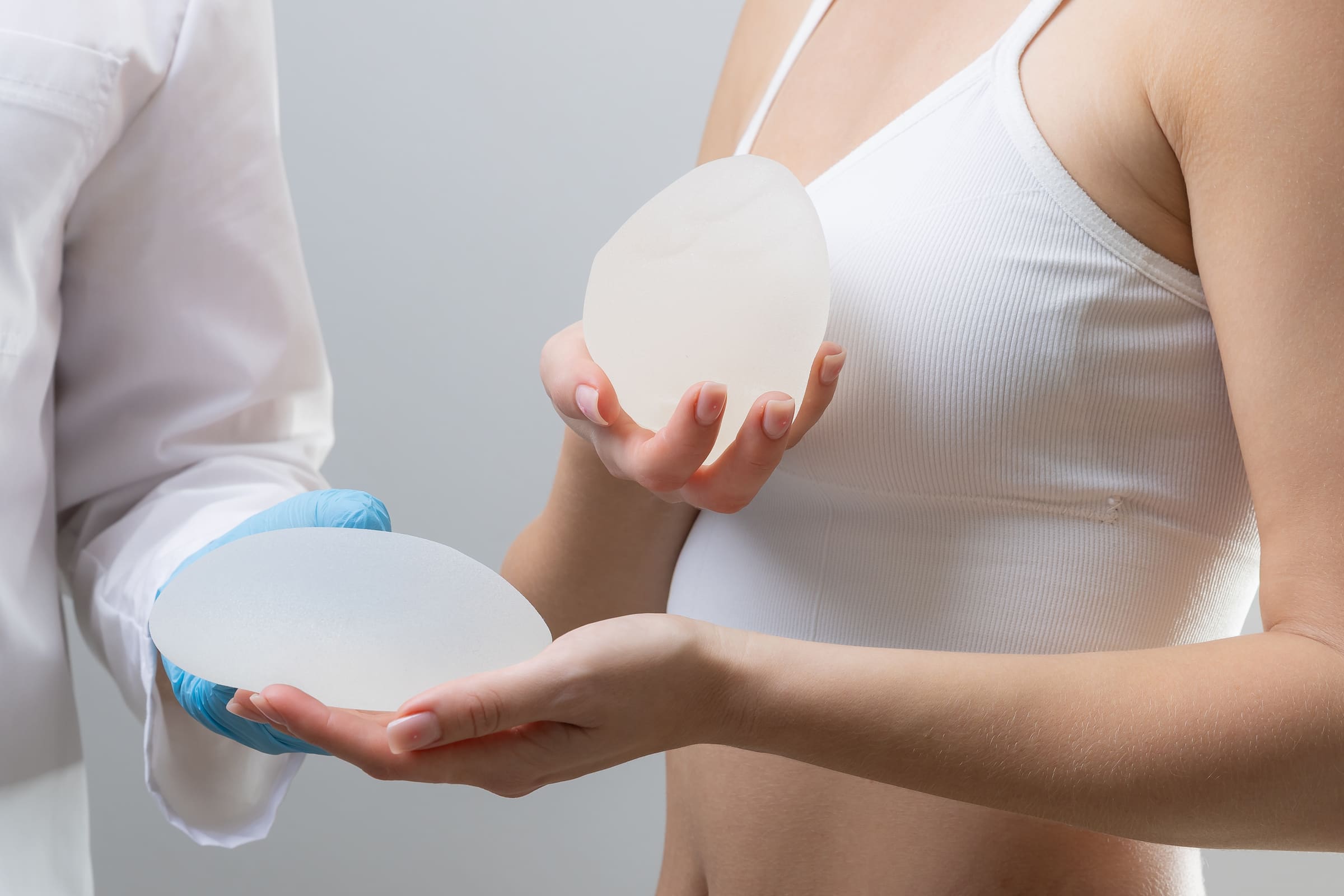 A woman in a white camisole holding a breast implant while a clinician in a white coat holds a second one beside her