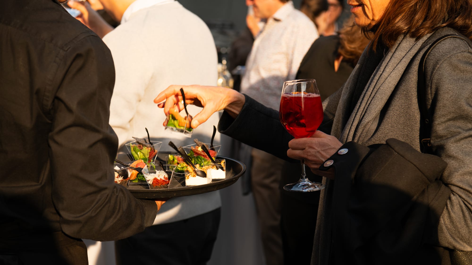 Guests at an evening reception: a server offers a tray of canapés while a guest reaches for one, another holding a glass of sparkling red drink, in warm low light.